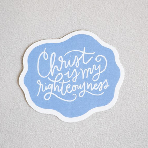 A die-cut sticker with a light blue background and white border, featuring the hand-lettered quote 'Christ loves my righteousness' in white script, shown on a neutral textured gray background.