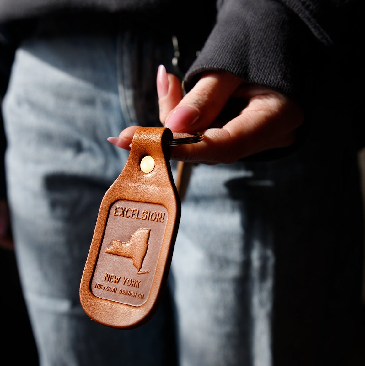 Brown leather keychain with Excelsior New York logo held by a person.