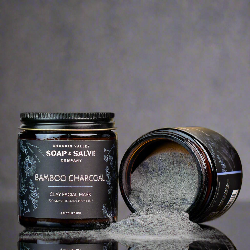Chagrin Valley Soap &amp; Salve Company Bamboo Charcoal Clay Facial Mask in a dark amber jar, with one jar open and its contents spilled onto a reflective surface against a gray background.