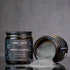 Chagrin Valley Soap & Salve Company Bamboo Charcoal Clay Facial Mask in a dark amber jar, with one jar open and its contents spilled onto a reflective surface against a gray background.