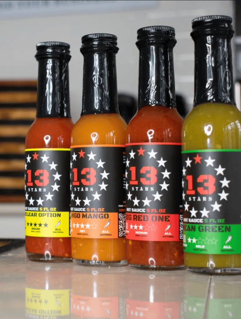 Four bottles of '13 Stars' hot sauce with different flavors on a reflective surface.