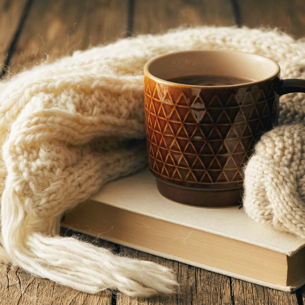 A warm brown ceramic mug with a geometric triangle pattern sits on a book, wrapped in a chunky cream knit scarf on a rustic wooden table.