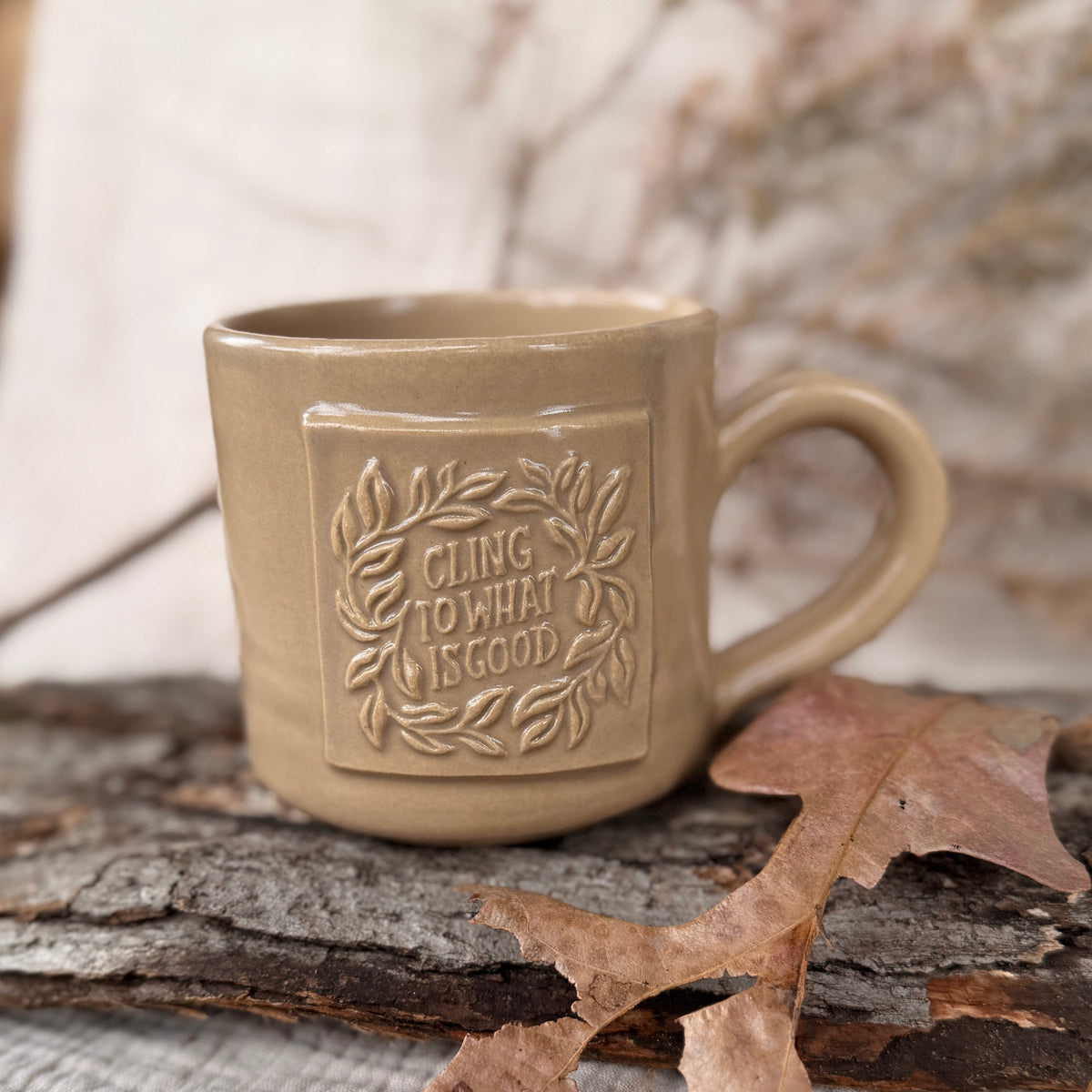 A beige ceramic mug with a handle and embossed floral border design featuring the text &