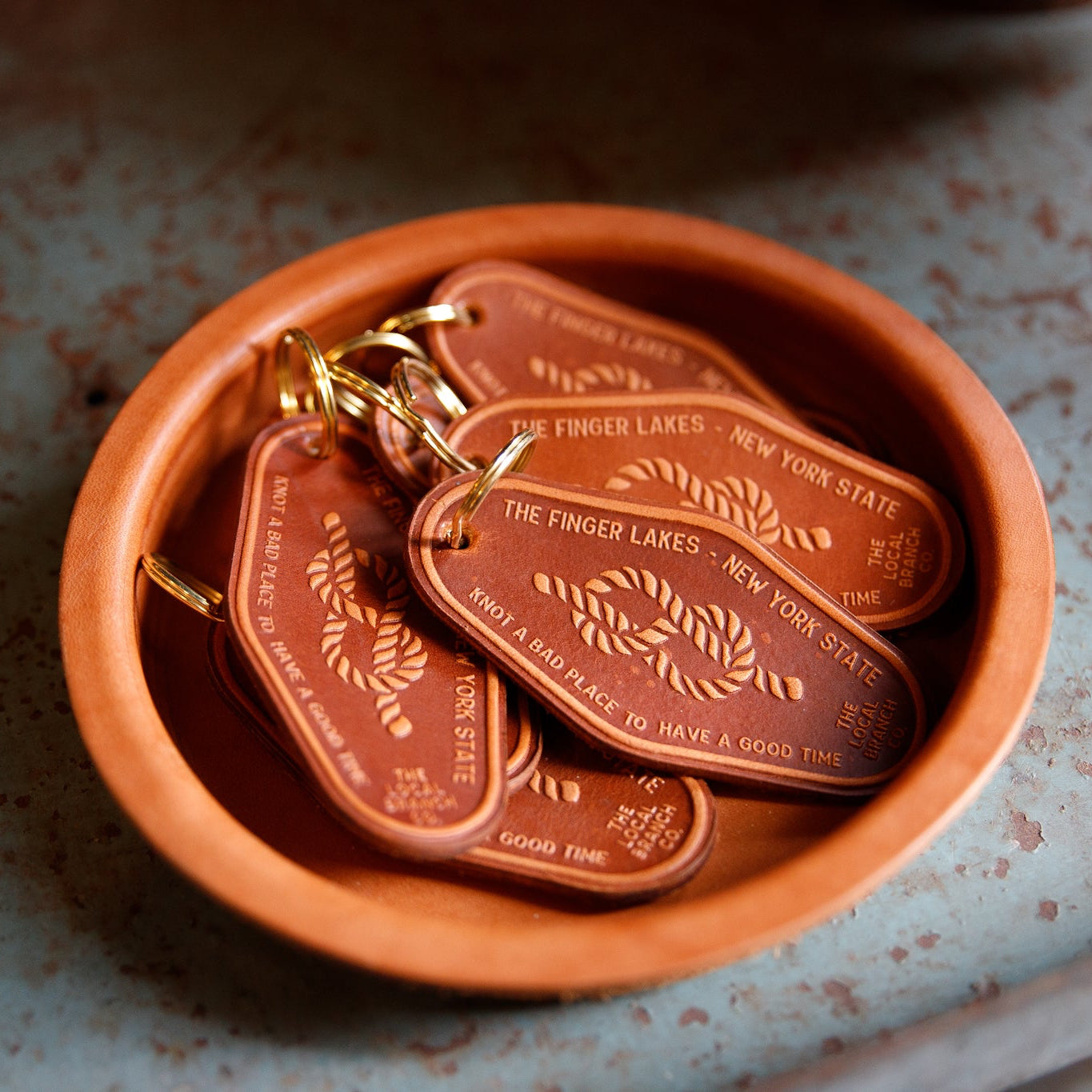 Brown leather keychains with gold text in a terracotta dish on a textured surface