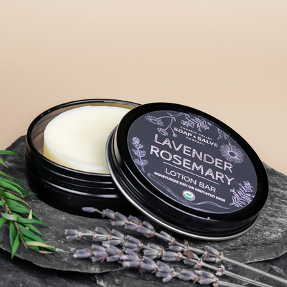 An open round black tin of Lavender Rosemary lotion bar with the lid leaning beside it, styled on a dark stone surface with lavender sprigs, a wooden bowl, and a soap dish.