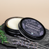 An open round black tin of Lavender Rosemary lotion bar with the lid leaning beside it, styled on a dark stone surface with lavender sprigs, a wooden bowl, and a soap dish.
