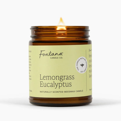 An amber glass jar candle with a lit flame, featuring a pale yellow-green label that reads &