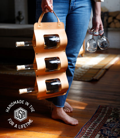 Person holding a wooden bottle holder with bottles and glasses indoors.