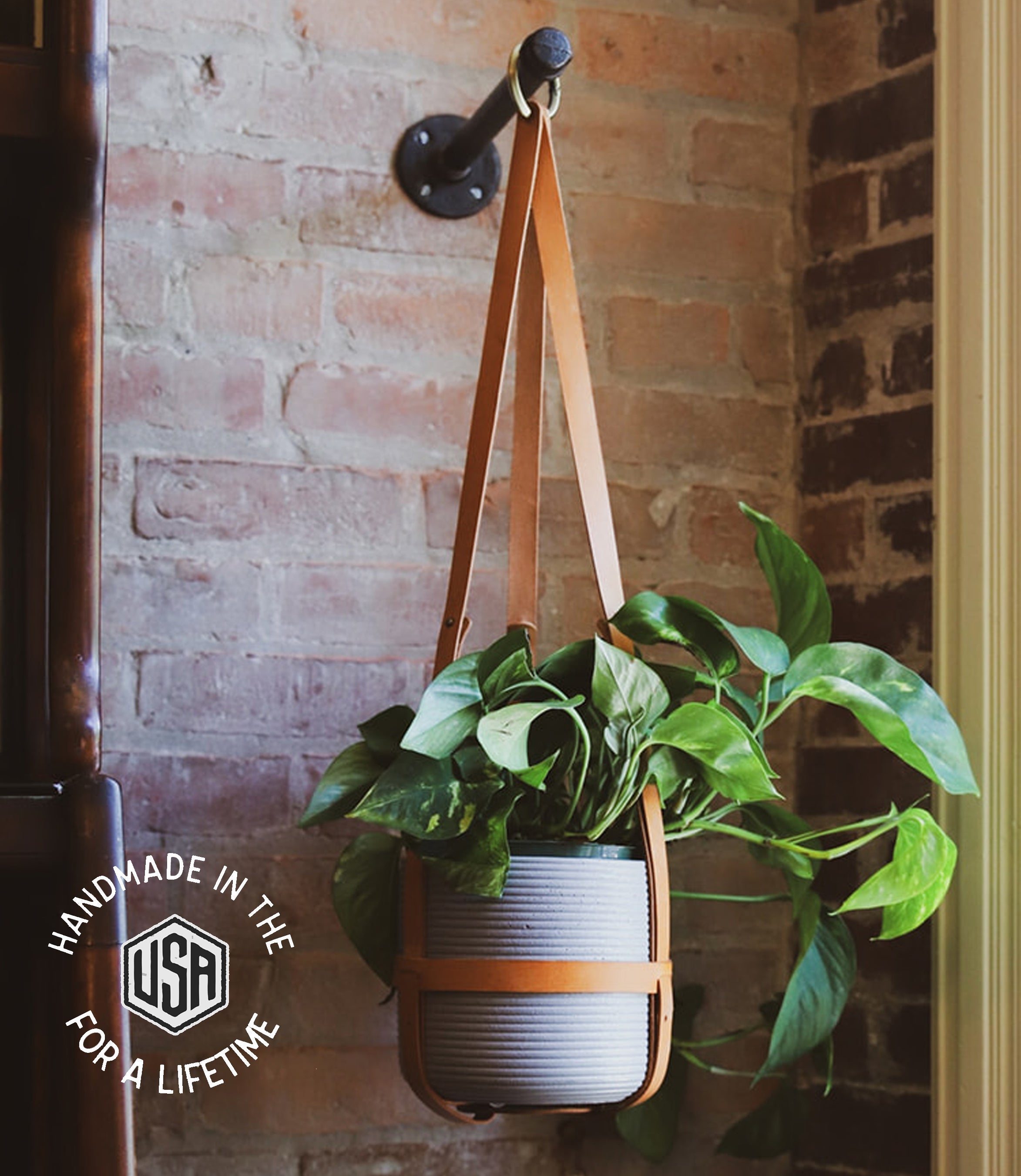 Hanging plant holder with leather strap against a brick wall, featuring &