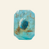A faceted turquoise-blue crystal or gemstone with a central rougher blue inclusion and scattered brownish inclusions, photographed upright on a clean light gray to white studio background.