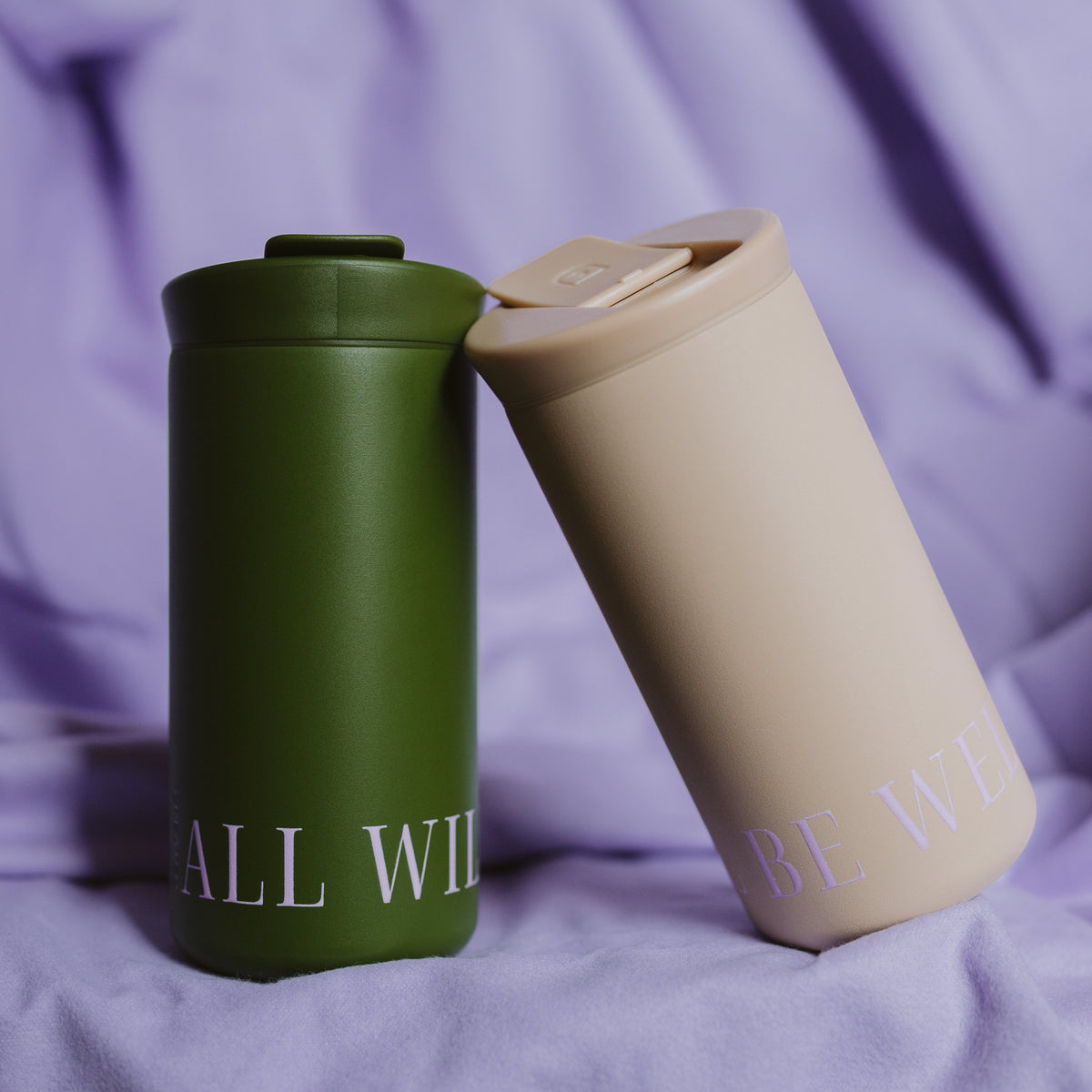 Two matte insulated tumblers resting on draped lavender fabric. One tumbler is olive green with white text reading &