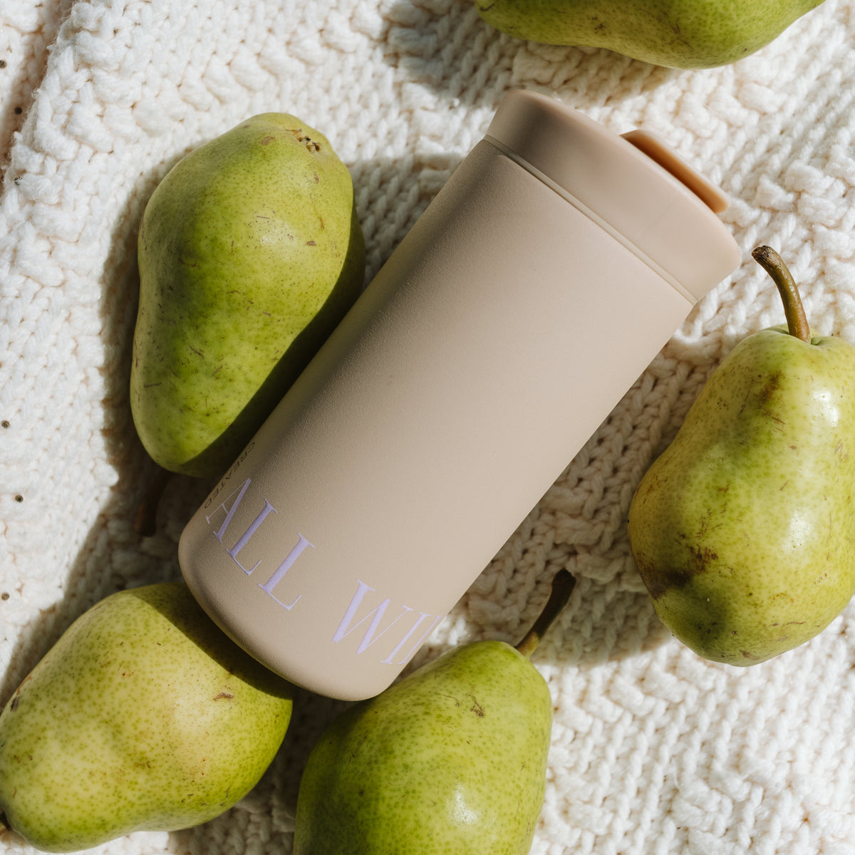 A matte beige insulated tumbler with a matching lid and a small orange accent on the spout, placed on a textured cream knit blanket and surrounded by green pears.