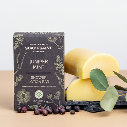 Chagrin Valley Soap &amp; Salve Company Juniper Mint Shower Lotion Bar packaging box shown next to two yellow bars of soap on a dark slate surface with green leaves and berries.