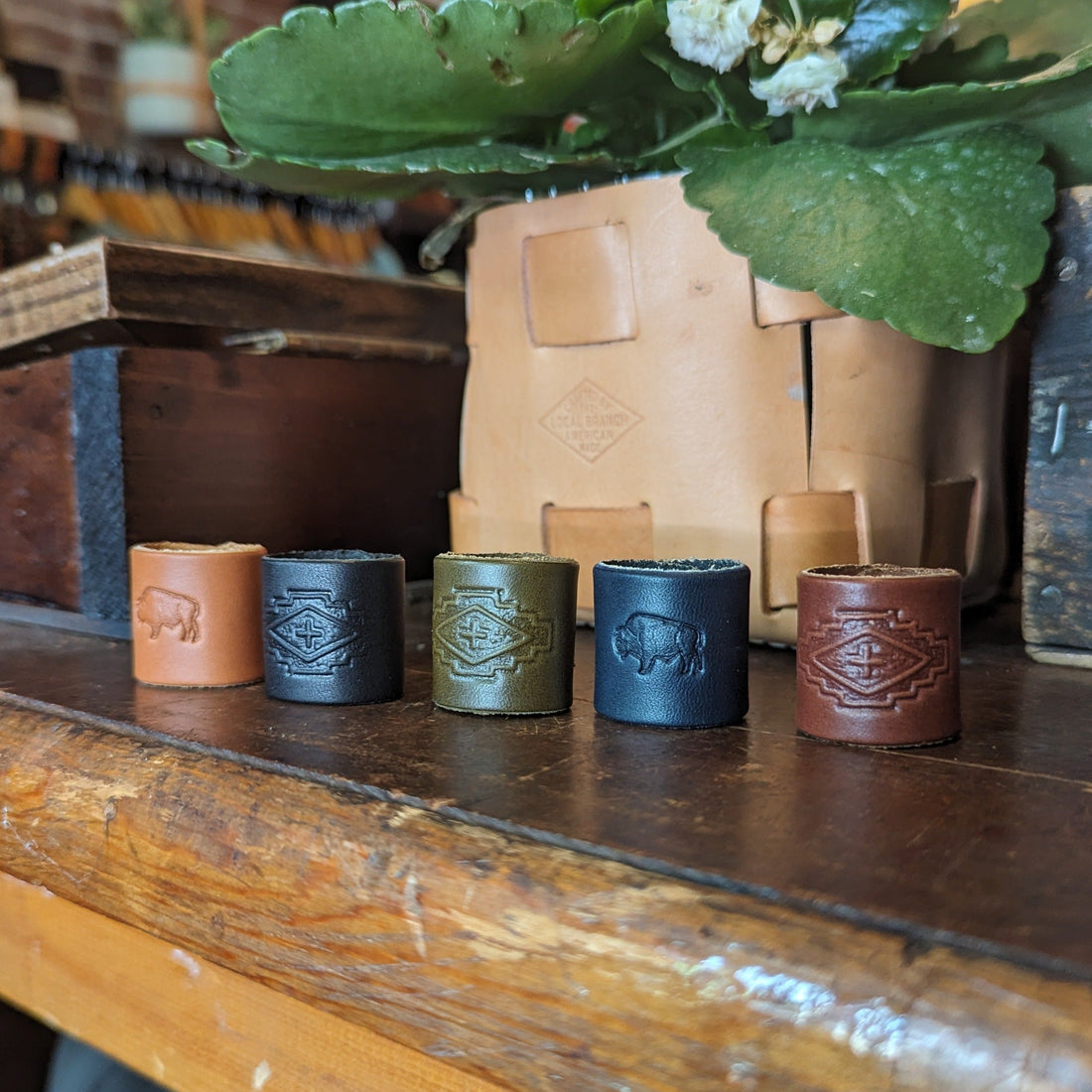 Set of five leather cups with embossed designs on a wooden surface, with a plant and wooden box in the background.