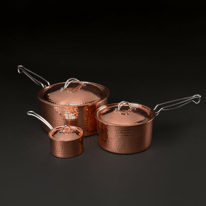 Three hammered copper saucepans with matching lids in different sizes, arranged on a dark studio background.