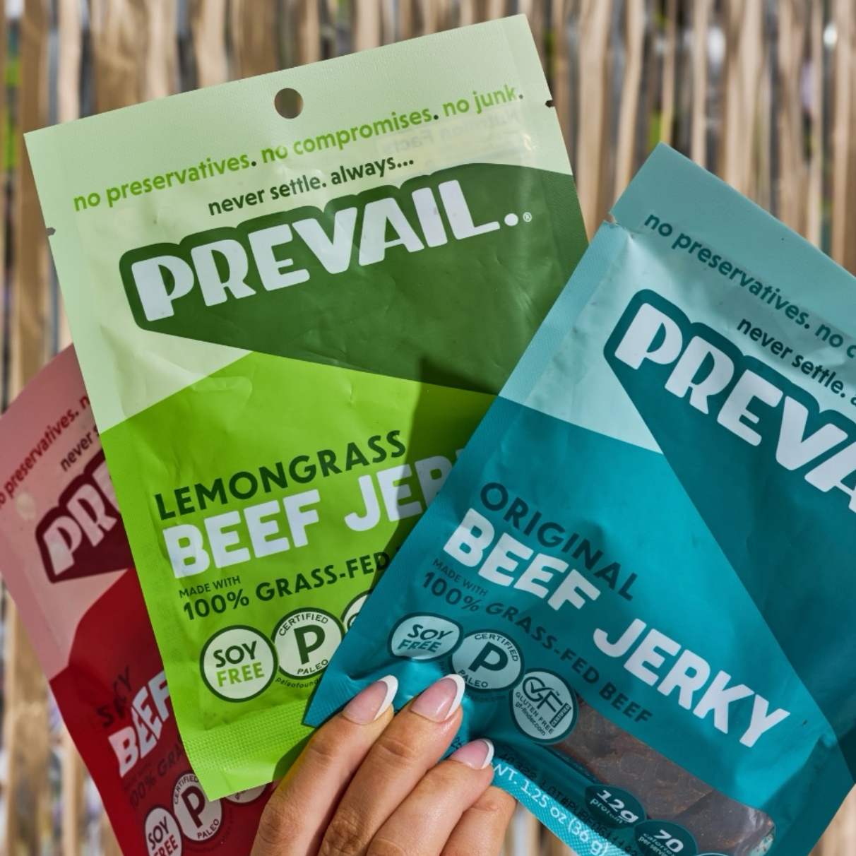 A hand holding three bags of Prevail beef jerky against a background of dried palm leaves. The bags are in green, blue, and red packaging, labeled 'Lemongrass Beef Jerky', 'Original Beef Jerky', and 'Beef Jerky' respectively.
