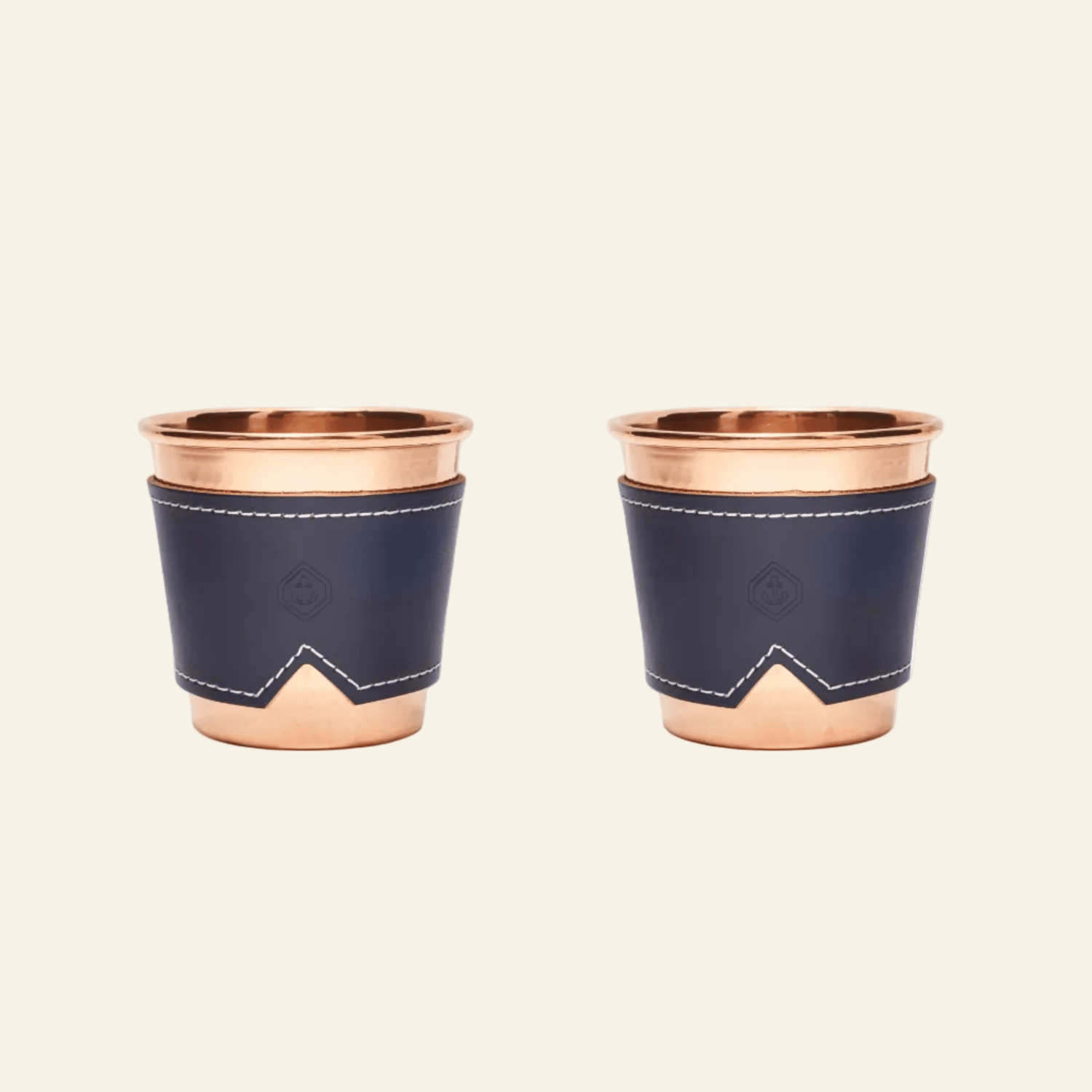 Two copper cups with navy blue leather sleeves on a beige background