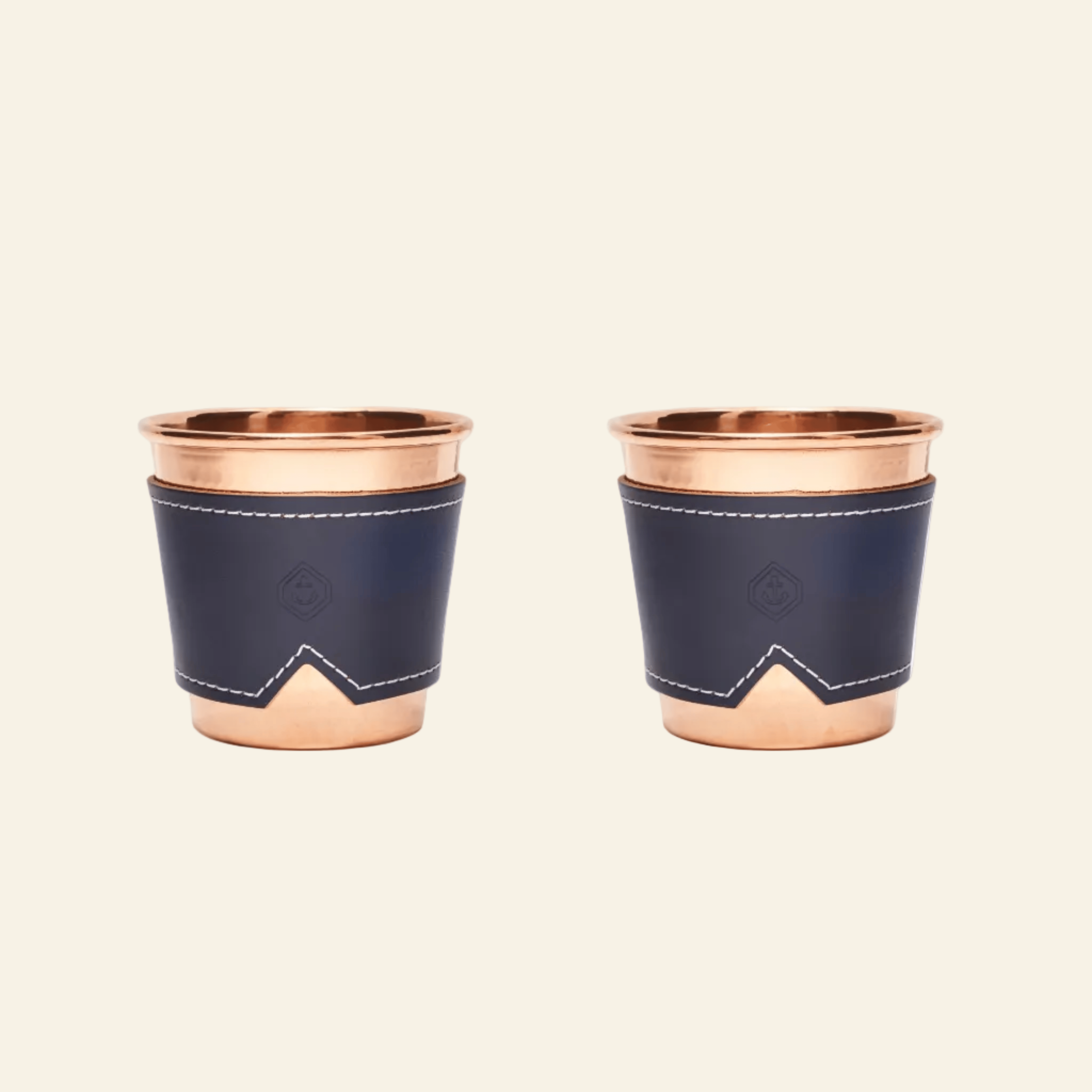 Two copper cups with navy blue leather sleeves on a beige background