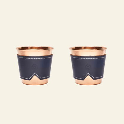 Two copper cups with navy blue leather sleeves on a beige background