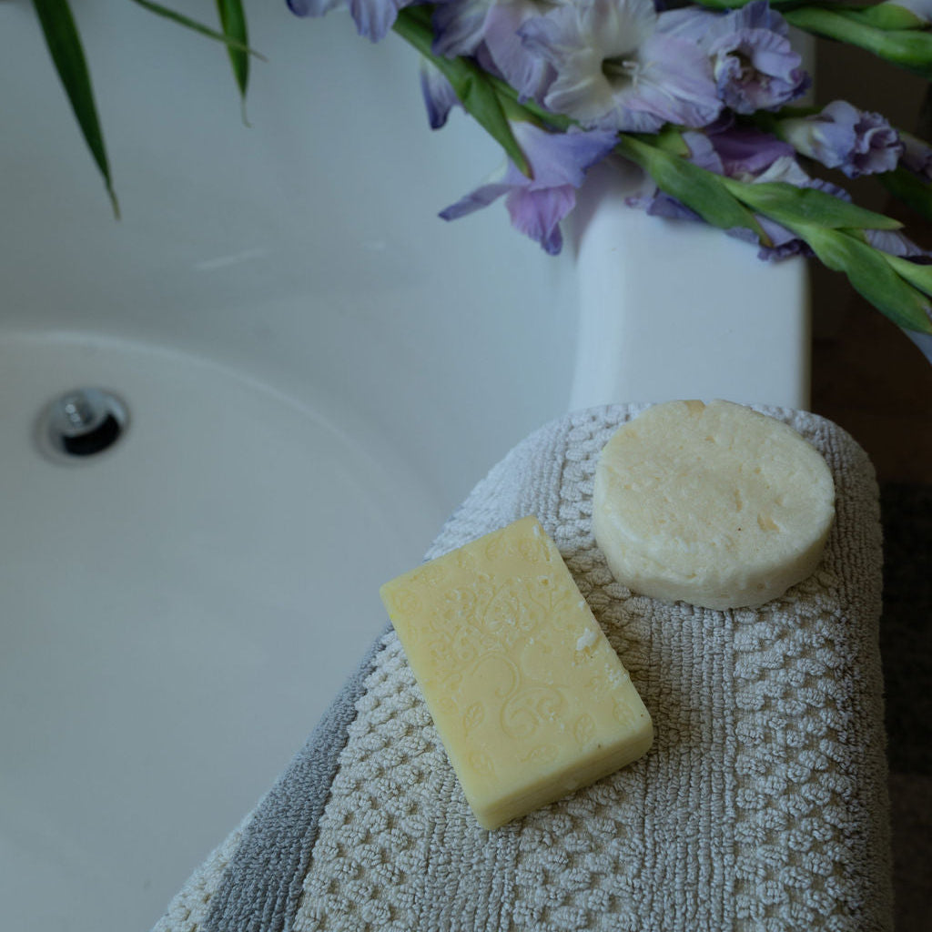 A close-up of a textured beige towel draped over a white sink, with two round bars of soap resting on it and purple gladiolus flowers in the background.