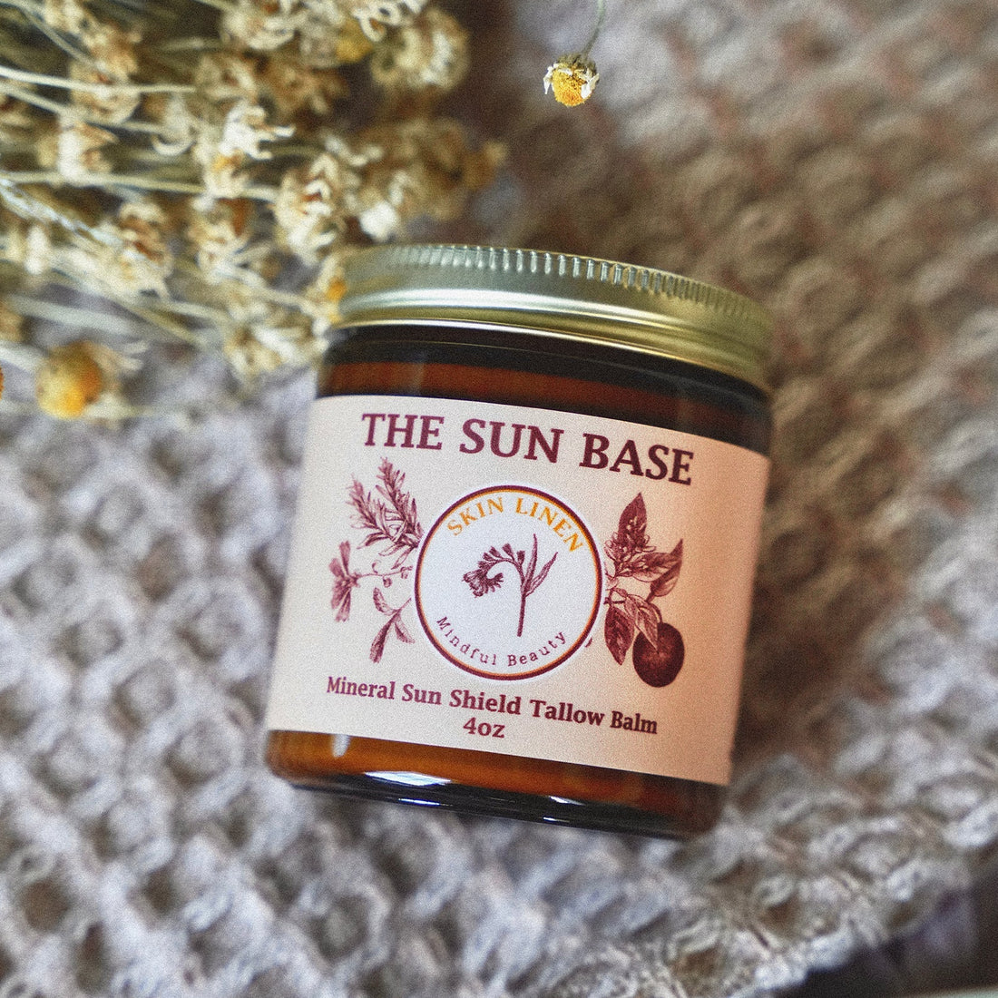A small amber glass jar of The Sun Base Skin Line Mineral Sun Shield Tallow Balm with a gold lid and cream label, styled on a textured beige blanket with dried flowers.