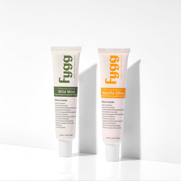 Two Fygg toothpaste tubes standing upright on a white surface against a white wall, with one tube in Wild Mint and one in Vanilla Aldehydes.