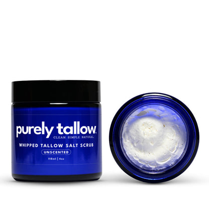 A dark blue jar of Purely Tallow Whipped Tallow Salt Scrub with a black lid, shown next to its open lid revealing white whipped cream inside, on a white background.