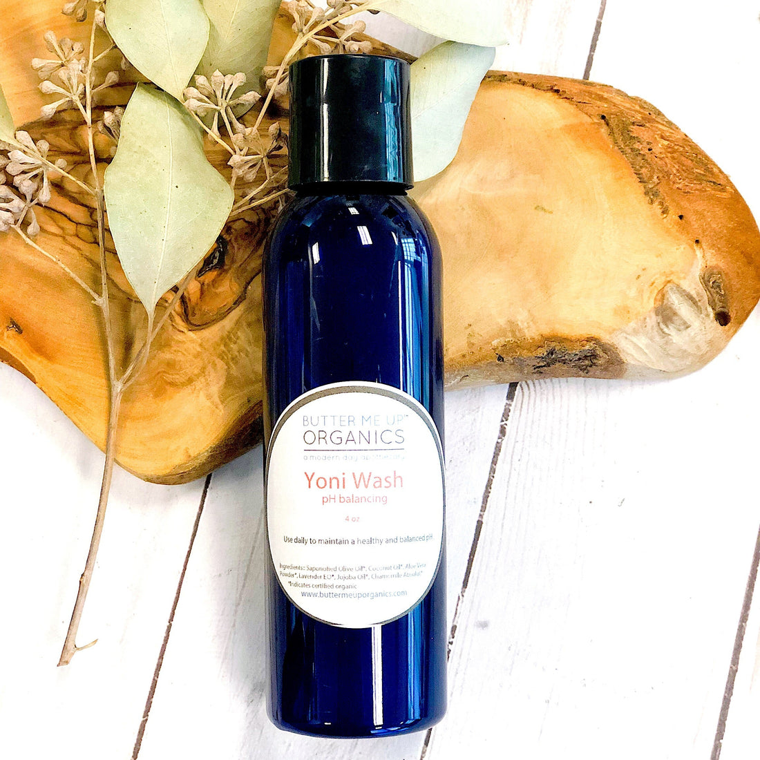 Blue bottle of Yoni Wash with a white label on a wooden surface with dried leaves.