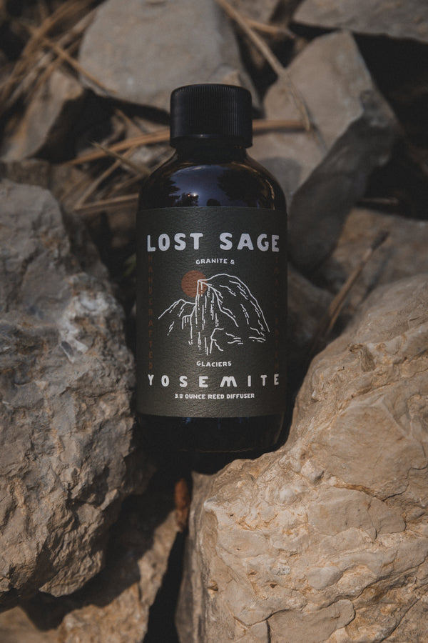 A dark amber glass bottle of Lost Sage Granite 5 Yerba Mate, featuring a black label with white text and mountain graphics, nestled between natural gray rocks.