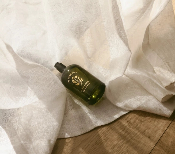 A dark green glass bottle with gold lettering lies on draped white fabric over a wooden surface. The bottle appears to be the product being sold, photographed in a styled flat-lay setup.