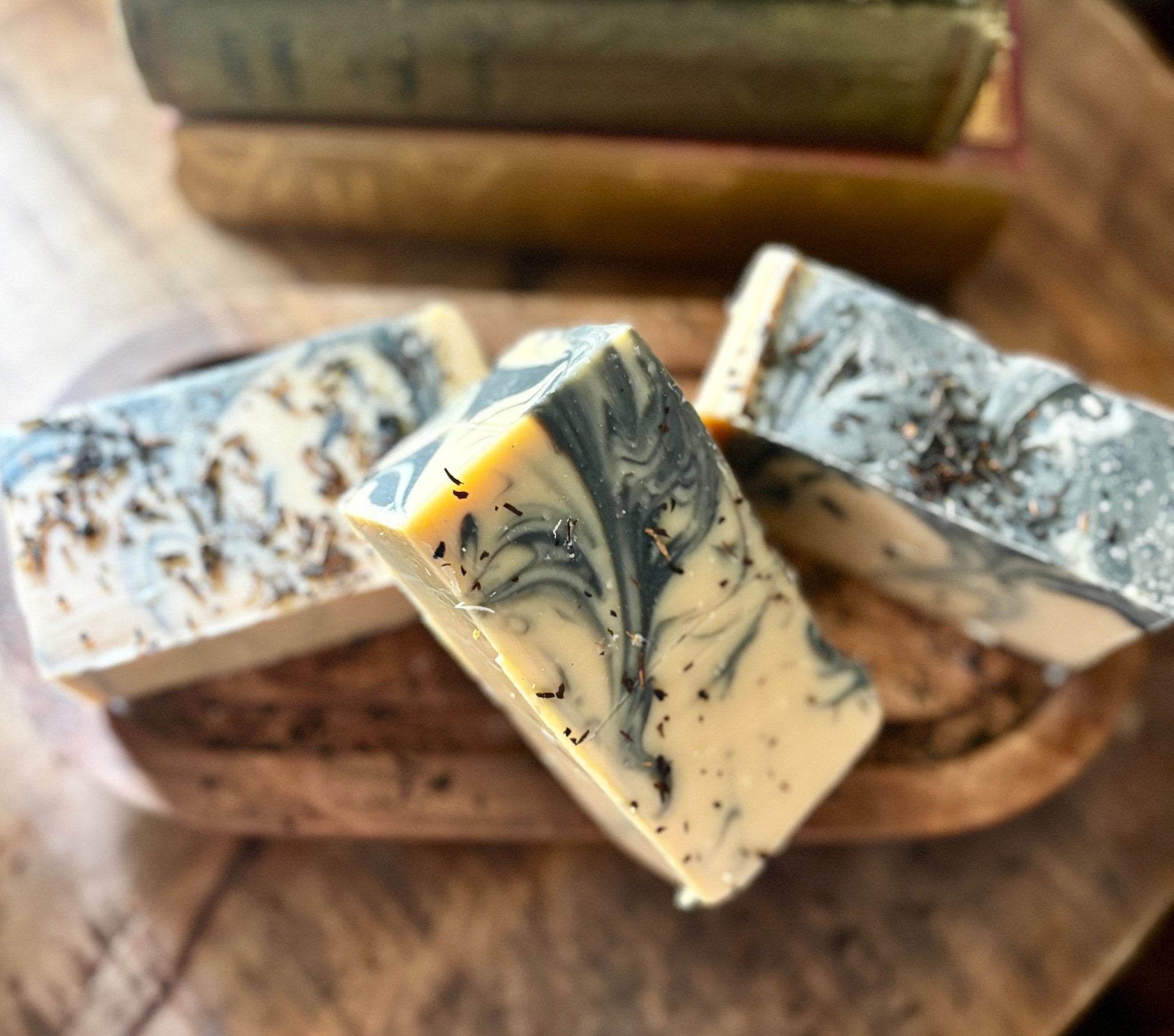 Three artisanal soap bars with a marbled cream, gray, and black swirl pattern, resting on a rustic wooden tray with vintage books in the background.