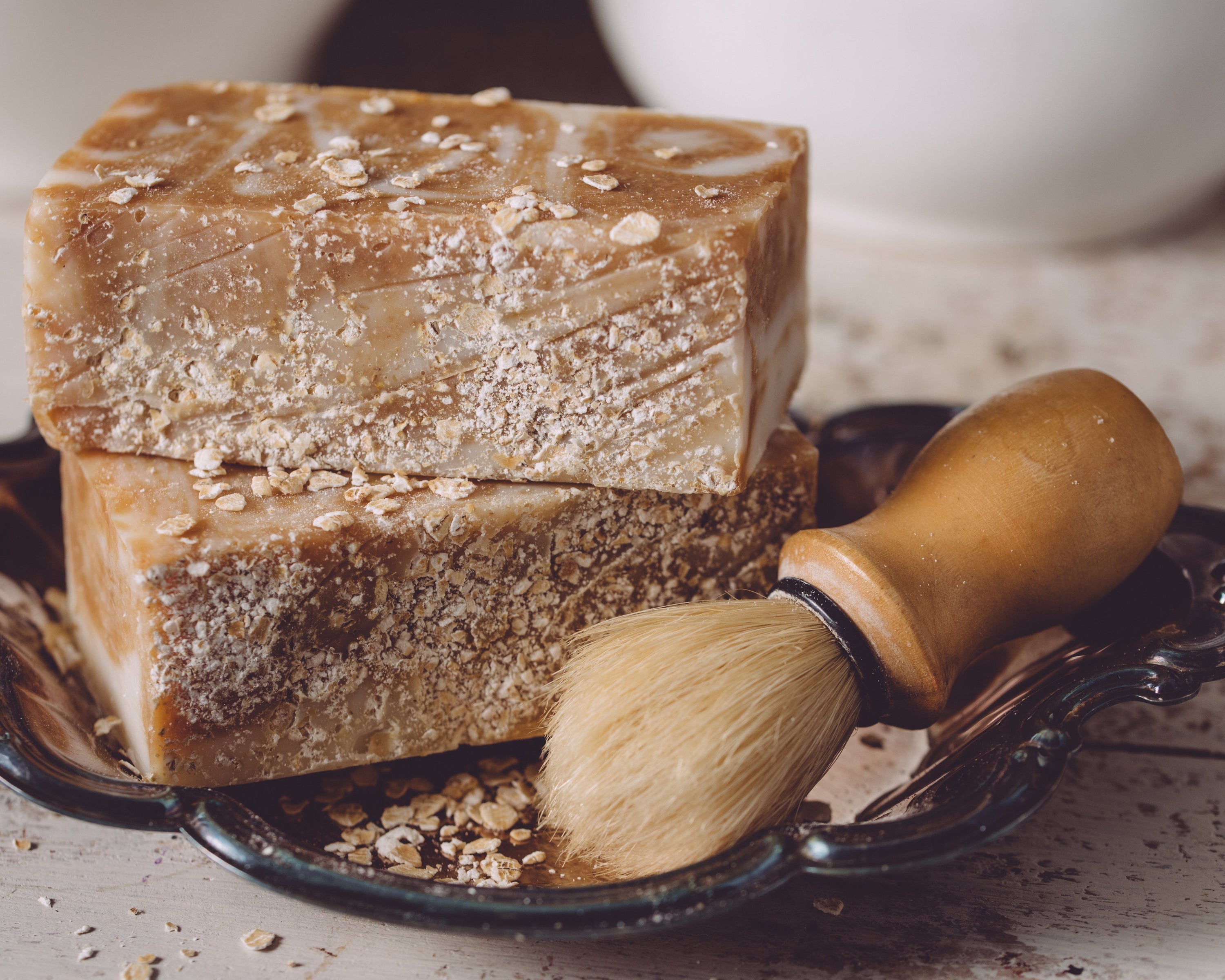 Two bars of brown soap with oatmeal on a black dish, accompanied by a wooden-handled brush.
