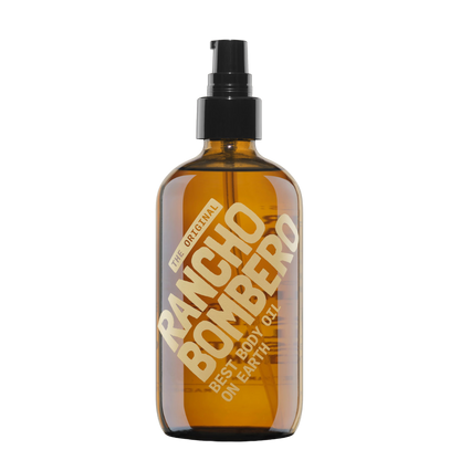 An amber glass body oil spray bottle with a black pump dispenser. The bottle features a large diagonal gold label that reads &