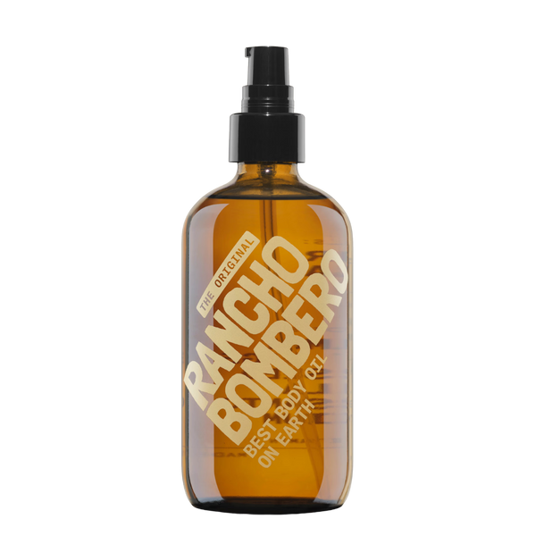 An amber glass body oil spray bottle with a black pump dispenser. The bottle features a large diagonal gold label that reads 'THE ORIGINAL RANCHO BOMBER BEST BODY OIL ON EARTH'.
