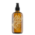 An amber glass body oil spray bottle with a black pump dispenser. The bottle features a large diagonal gold label that reads &