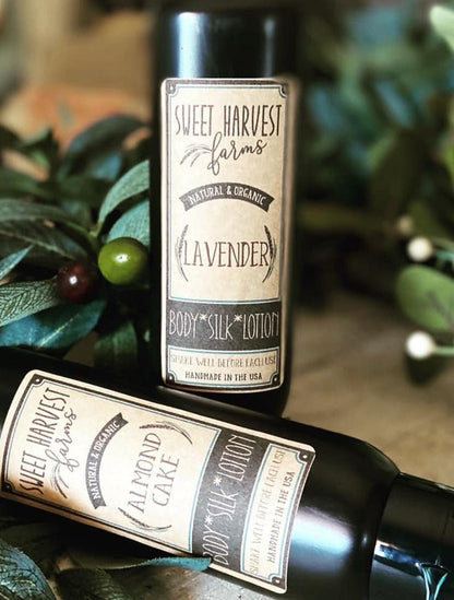 Two Sweet Harvest Farms body lotions in dark bottles, one standing upright and one lying down, surrounded by green leaves and small berries. The upright bottle is labeled Lavender, and the horizontal bottle is labeled Almond Cake.