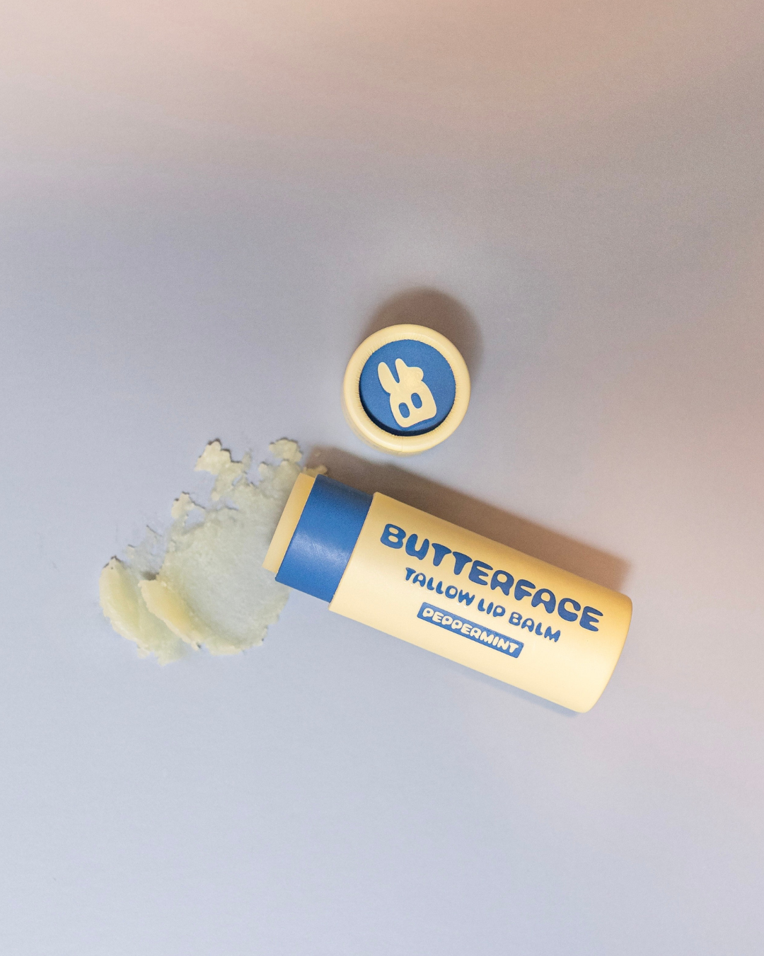 A tube of Butterface Tallow Lip Balm with its cap off, lying on its side with a smear of the pale yellow balm next to it, on a light gray background.
