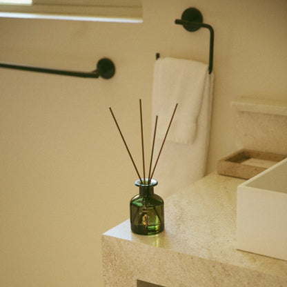 A green glass reed diffuser bottle with several dark reed sticks sits on a light beige stone bathroom counter next to a white sink basin.