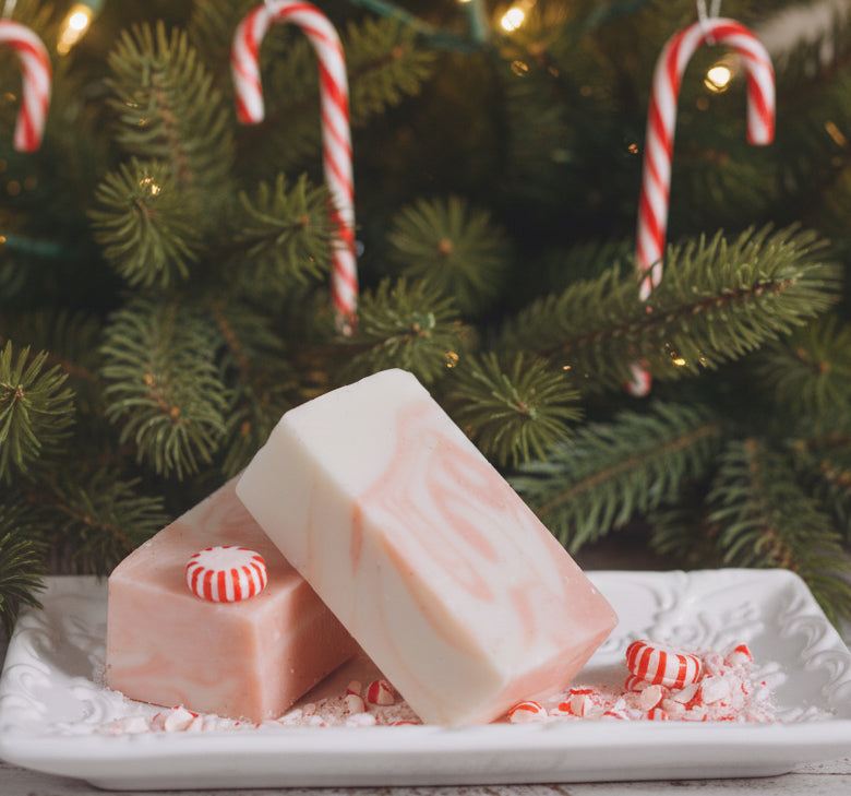 Two rectangular bars of soap with pink and white marbled patterns are arranged on a white decorative tray, surrounded by crushed candy cane pieces, with candy canes and evergreen branches in the background.