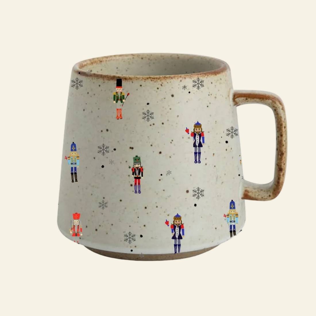 A speckled ceramic mug with a handle, decorated with small colorful nutcracker figures and snowflakes on a light gray background, set against a plain white studio backdrop.