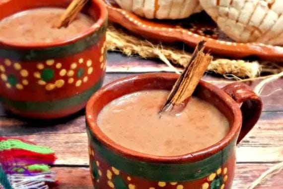 Two hand-painted Mexican clay cups filled with a brown hot chocolate or cocoa drink, garnished with cinnamon sticks, set on a rustic wooden table with traditional woven hats in the background.