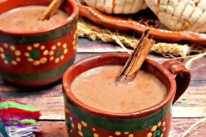 Two hand-painted Mexican clay cups filled with a brown hot chocolate or cocoa drink, garnished with cinnamon sticks, set on a rustic wooden table with traditional woven hats in the background.