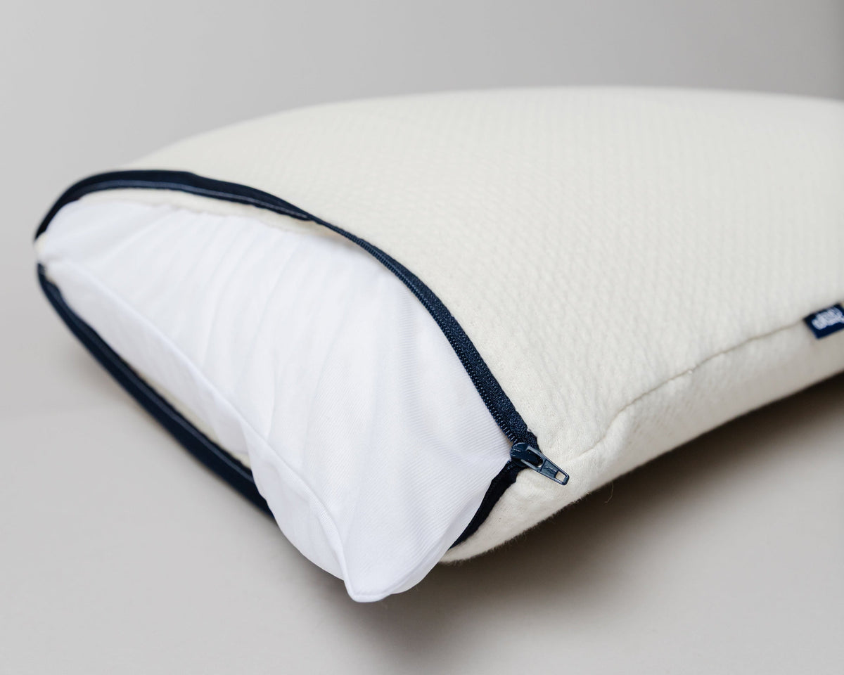 A close-up of a white pillow with a textured cover, partially unzipped to reveal a white inner pillow, finished with dark navy blue piping.