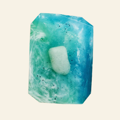 A hand holding a faceted blue-green crystal with a pale green stone embedded in the center, against a softly blurred light background.