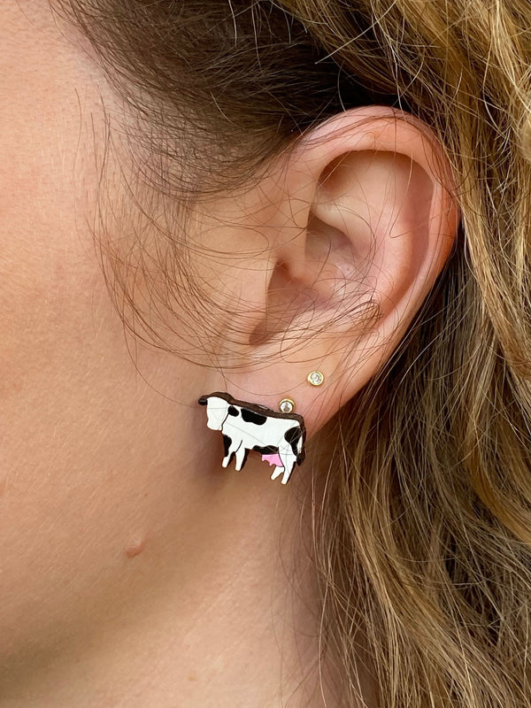 Two black and white cow earrings with pink udders on a beige background