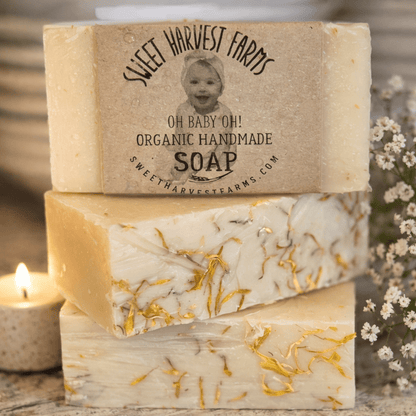 Three bars of soap with visible herbs, stacked on a wooden surface with a candle and flowers in the background.