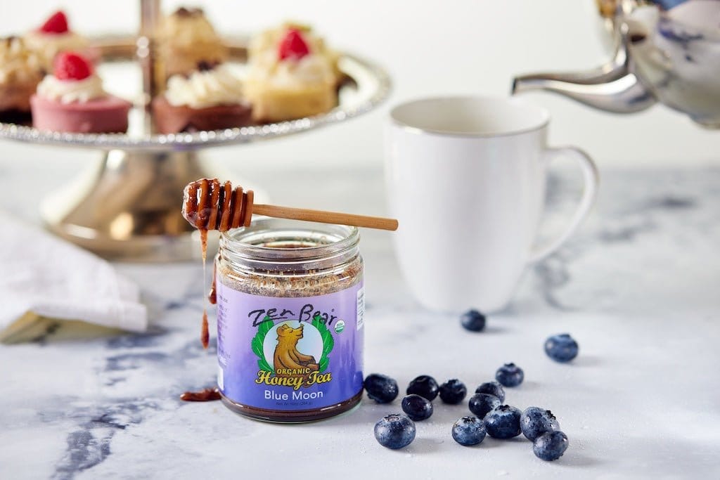 A jar of Bee-Blaze Honey Tea Blue Moon honey with a wooden honey dipper resting on the jar, set on a marble surface with blueberries, a white mug, and a teapot in the background.