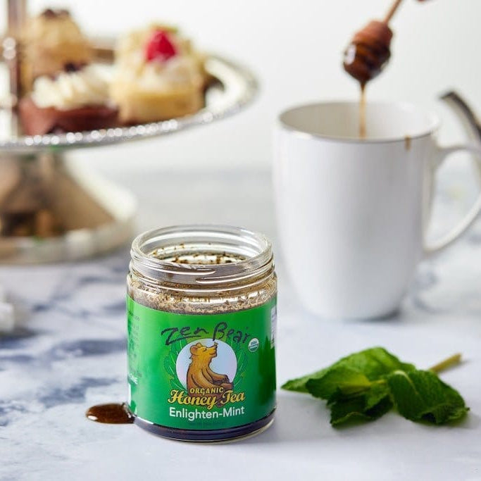 A small glass jar of Bee Gear Enlighten-Mint Honey Tea sits on a marble surface, with honey dripping from a wooden dipper into a white mug in the background. A green label with a bee logo is visible on the jar, and mint leaves and a cupcake stand are blurred in the scene.