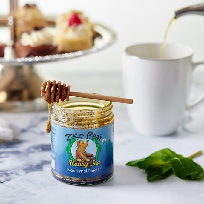 A jar of Tea Brise Handy Tea Nodumal Nectar with a honey dipper resting on top, set on a marble surface next to a white mug with tea being poured in and mint leaves, with cupcakes blurred in the background.