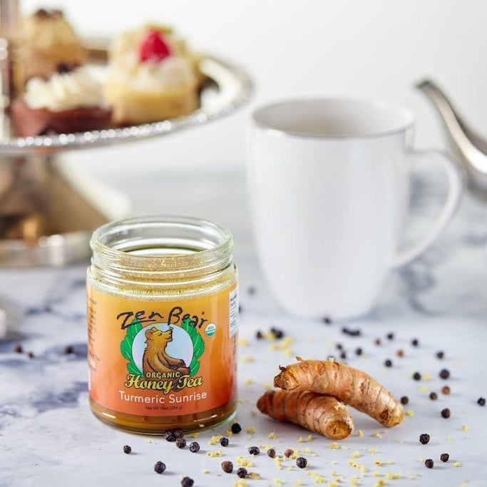 A glass jar of Turmeric Sunrise herbal tea by Tea Bites sits on a marble surface with a raw turmeric root, scattered peppercorns, a white mug, and a silver teapot in the background, alongside cupcakes on a stand.